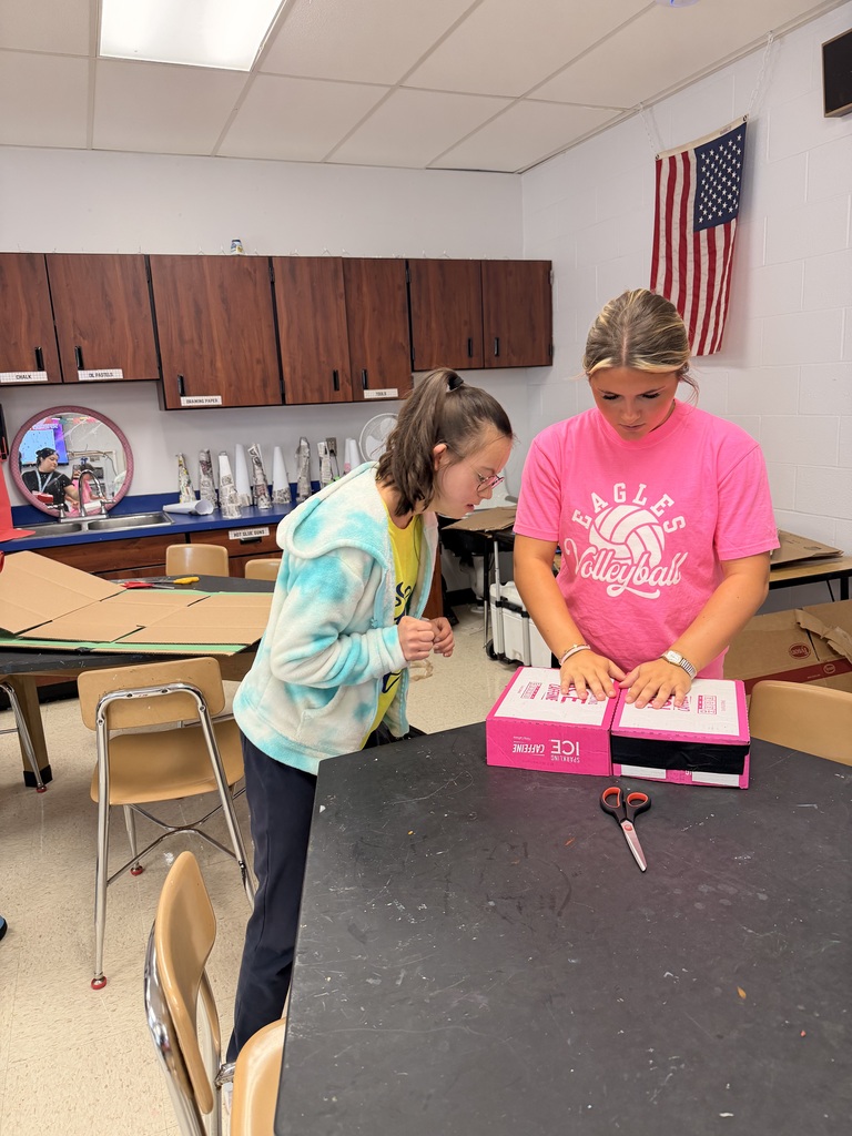 Two students work together on a pink cardboard box project at a classroom table, with scissors nearby.