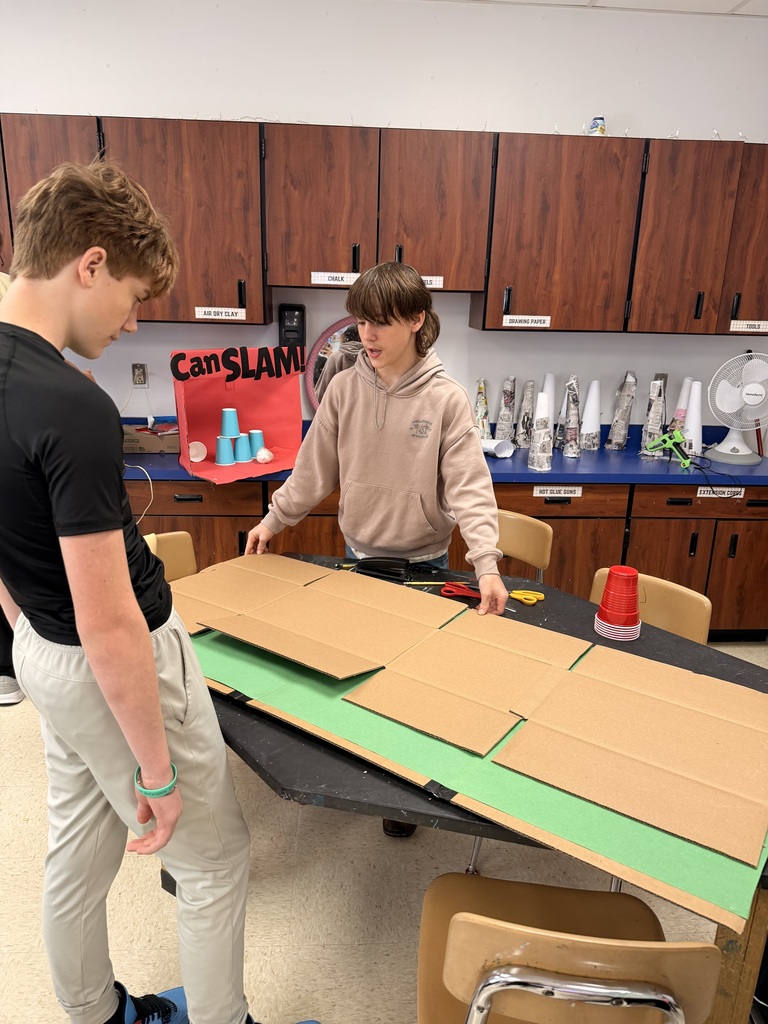 Two students arrange large cardboard panels over green material while building a classroom project.
