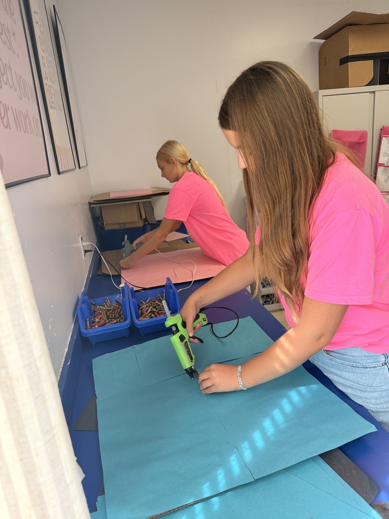 Two students in pink shirts use hot glue guns to assemble colorful cardboard pieces at a classroom worktable.