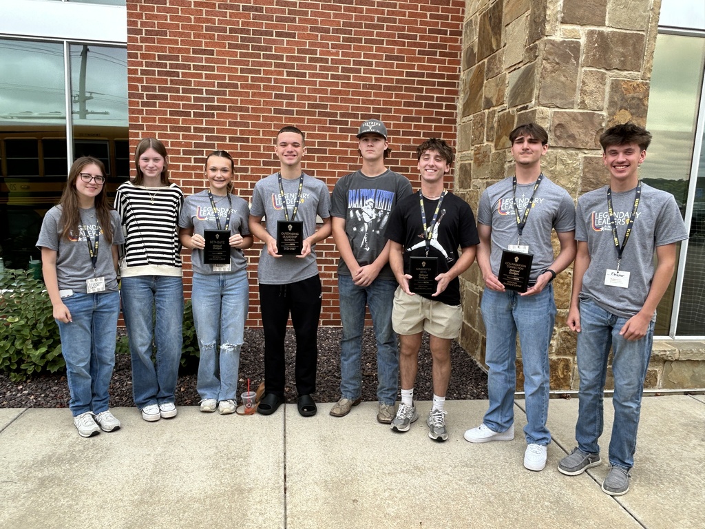 Eight students pose outside a brick building, with several holding leadership and achievement plaques.