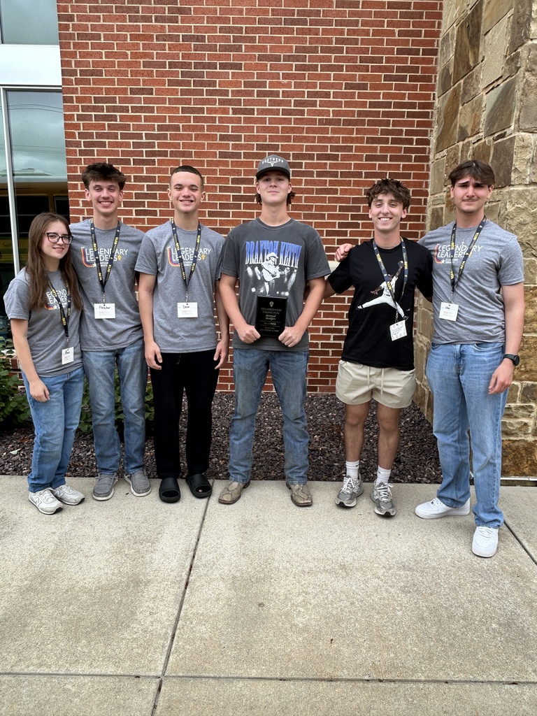 Six students pose outside a brick building, with the center student holding a collaboration award plaque.
