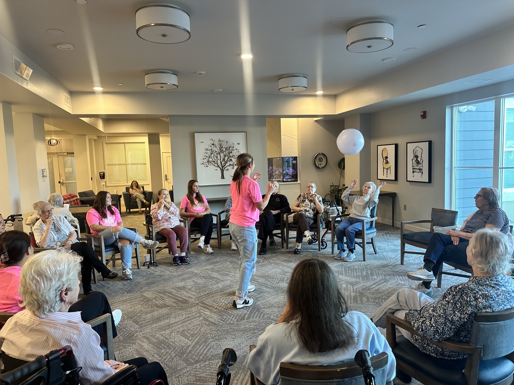A volleyball player leads seated older adults in a circle as a large white ball is tossed across the room.