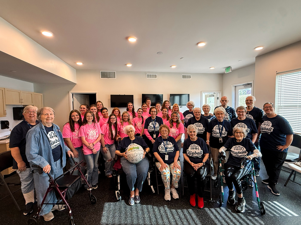 Volleyball players in bright pink shirts pose with older adults in navy shirts at a senior living community.