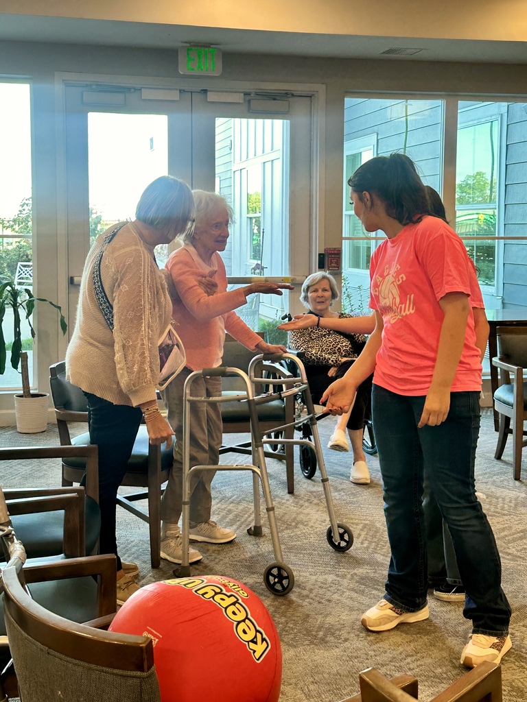 A volleyball player in a pink shirt engages with older women during a ball activity, with a walker nearby.
