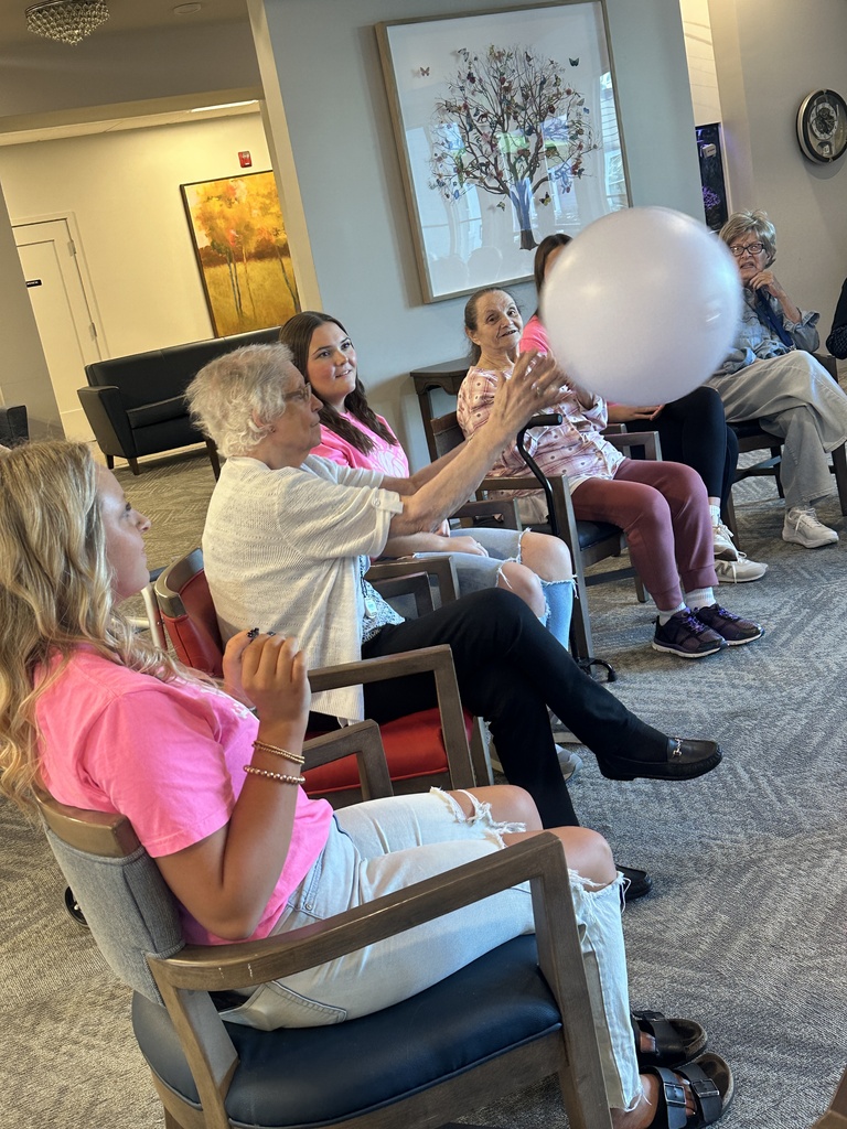 An older woman reaches for a large white ball while volleyball players and residents sit together in a lounge.