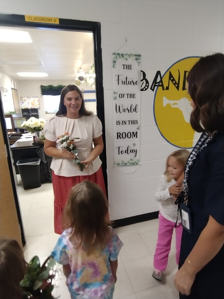 Young students deliver flowers to a smiling teacher standing in her classroom doorway.
