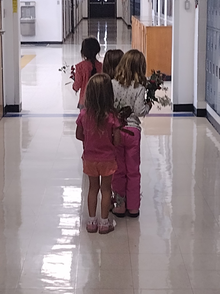 Four young students carry small bouquets of flowers as they walk together down a school hallway.