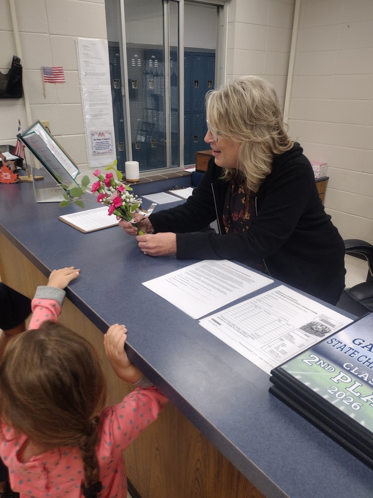 A school staff member smiles while holding a small bouquet presented by a young student at the office counter.
