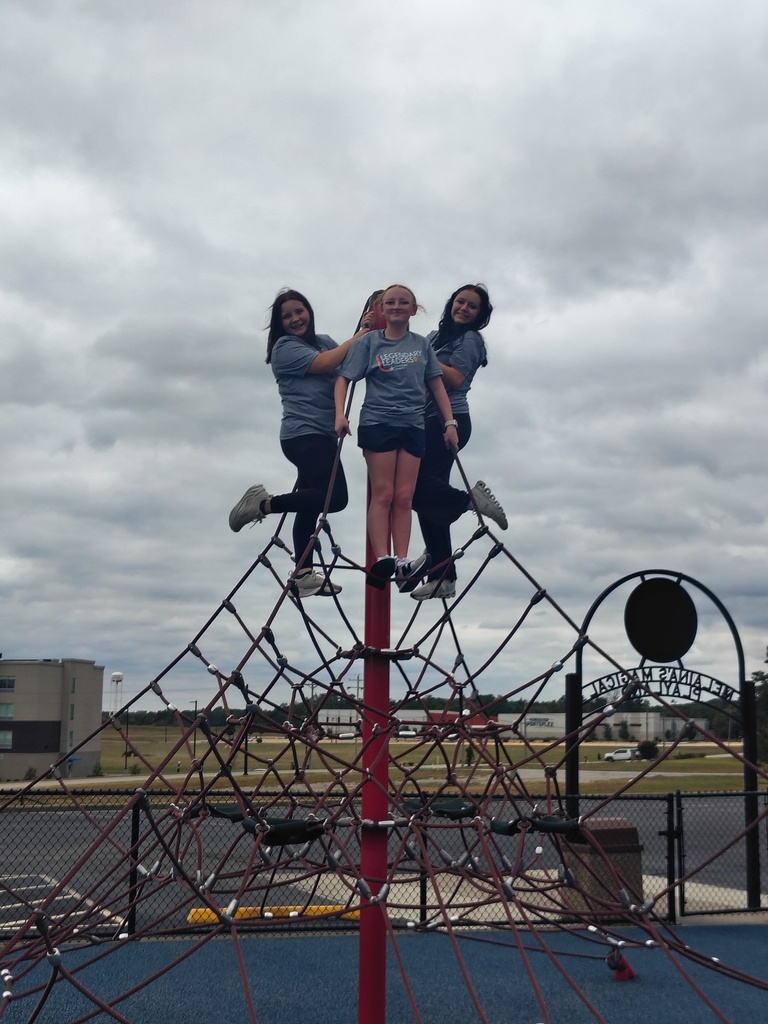 Three students in matching shirts pose atop a tall rope-climbing structure beneath a cloudy sky.