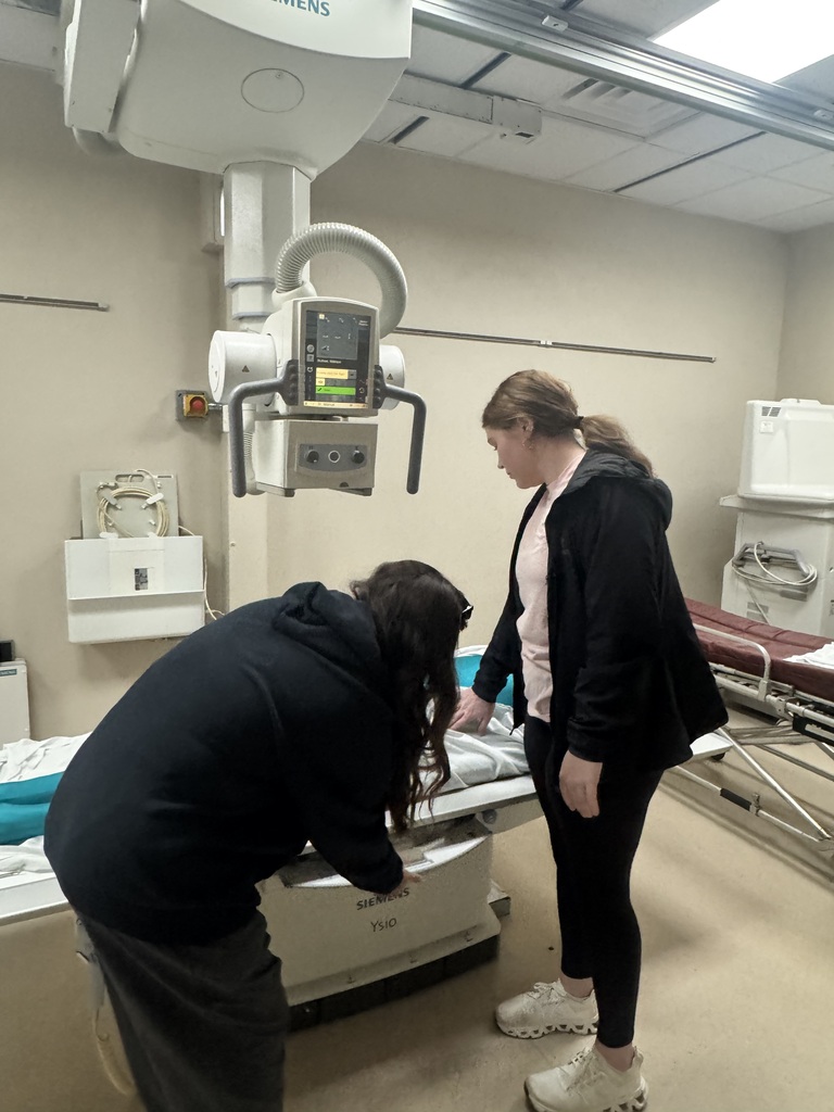 Two people practice positioning at an X-ray table beneath a large overhead imaging machine.