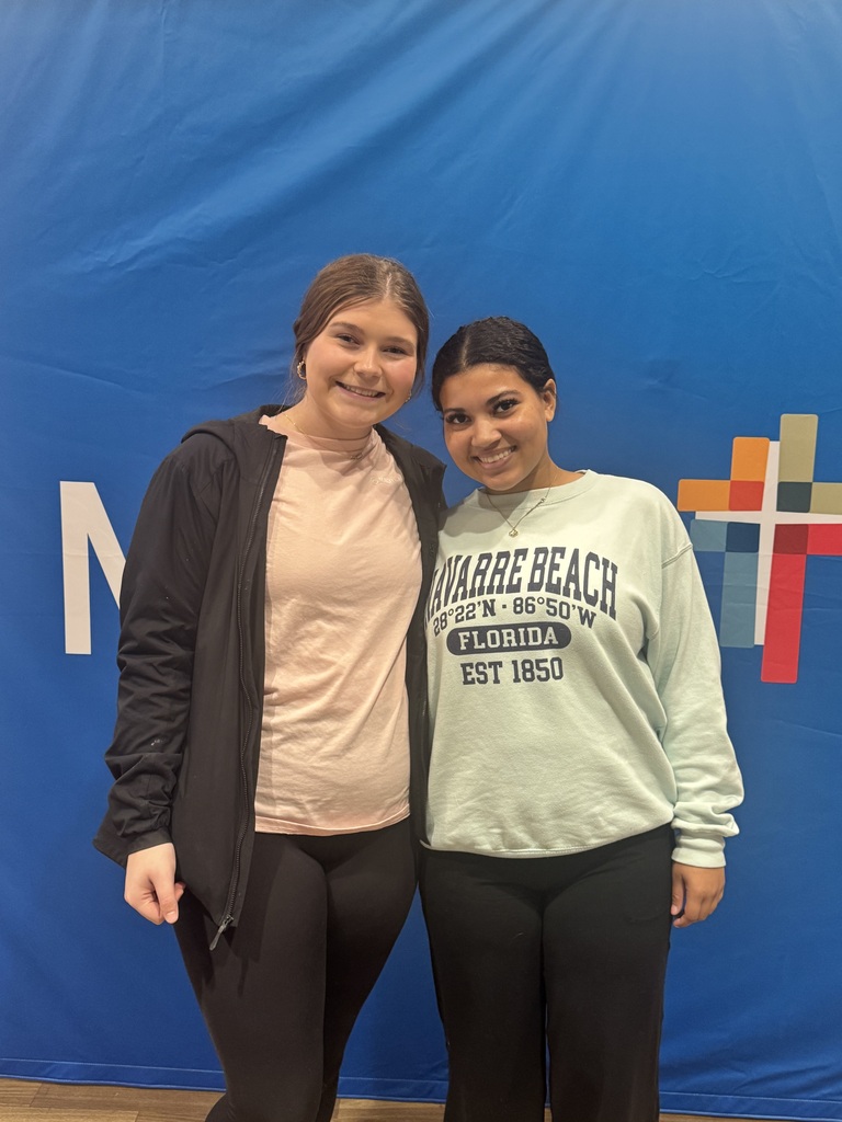 Two smiling women pose together in front of a bright blue backdrop with a colorful geometric logo.