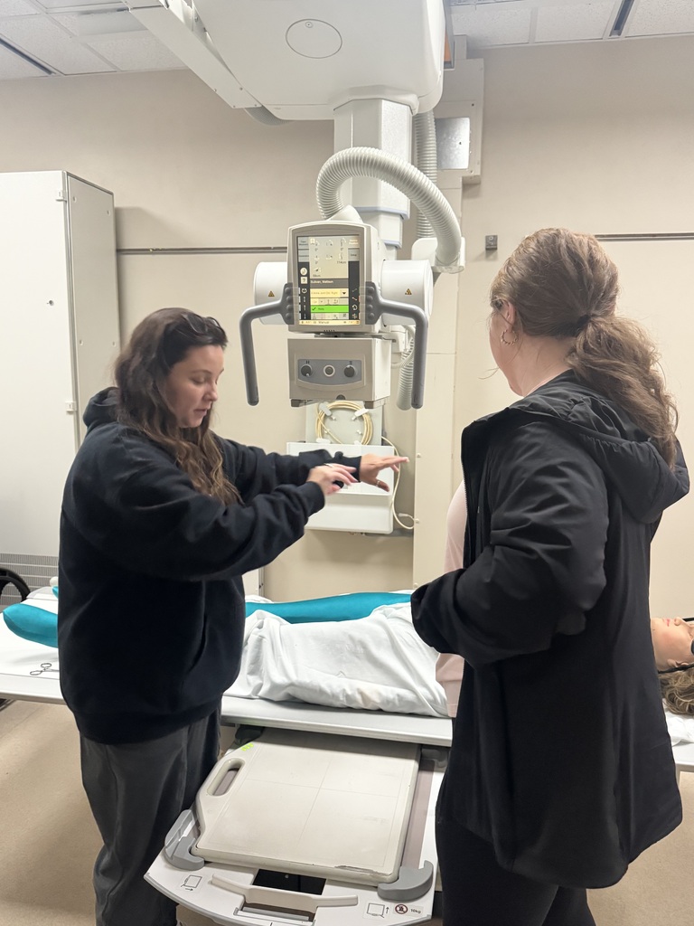 Two people stand beside an X-ray table as one demonstrates positioning near the imaging equipment.