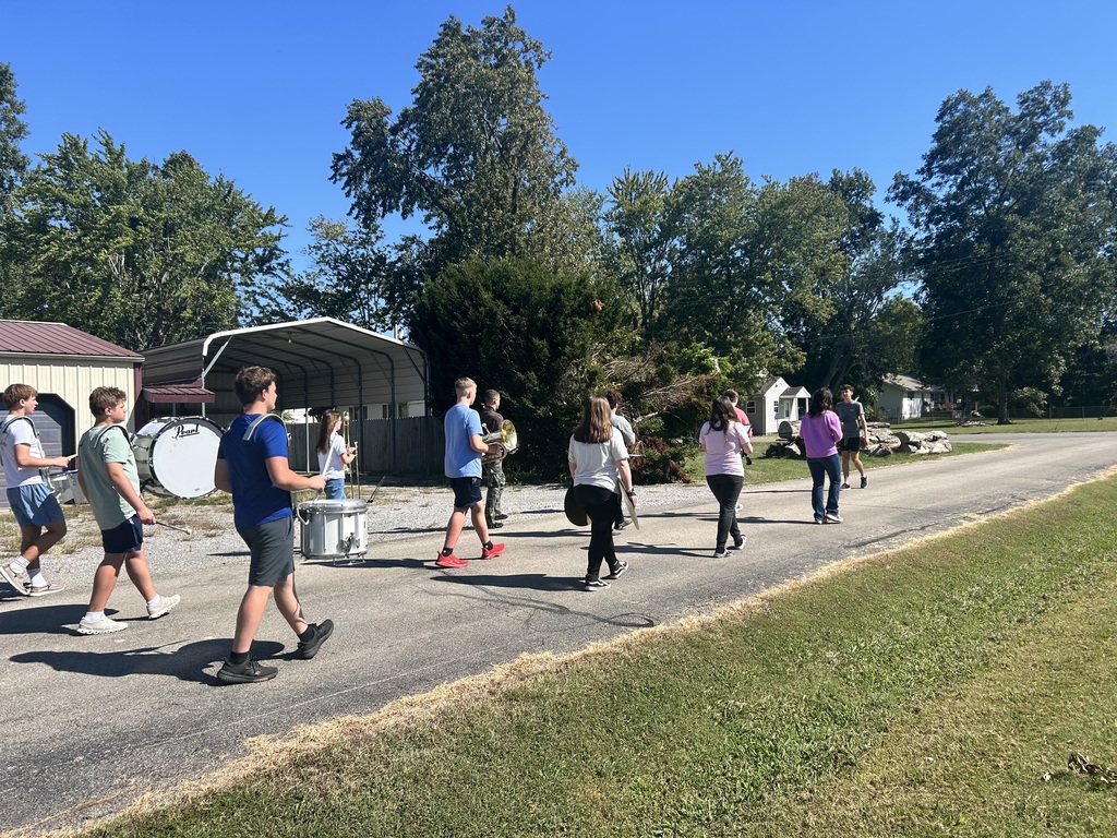 A group of students marches down a sunny residential street, carrying drums, cymbals, and brass instruments.