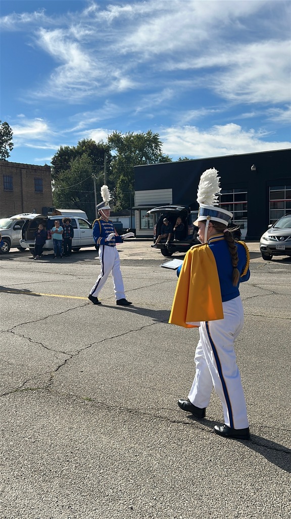 Two marching band members in blue, gold, and white uniforms walk along a sunlit street as spectators watch.
