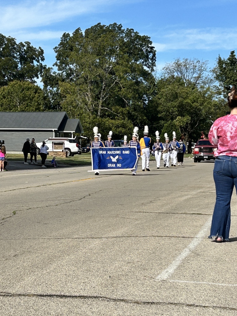 Marching band in blue-and-white uniforms carries an “Oran Marching Band” banner during a sunny street parade.