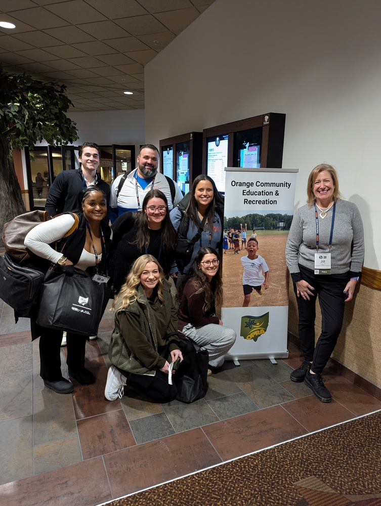 A group of people bunched around a banner with the words "Orange Community Education & Recreation" smiling at the camera