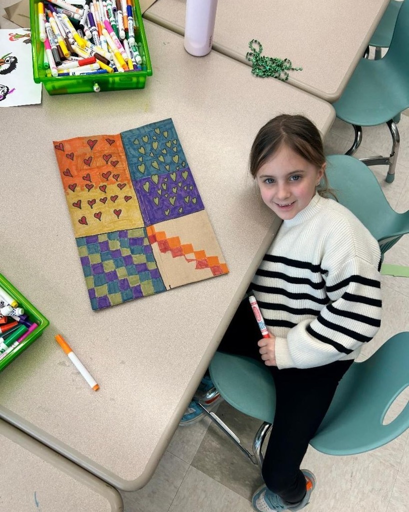 Young girl sitting at a desk with her artwork on the table, and she's smiling at the camera