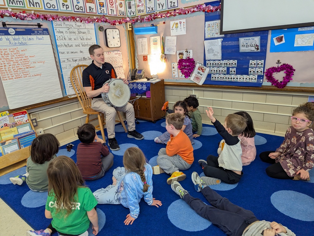 A group of preschool students sitting on a carpet, while a teacher holding a drum is teaching them about drums