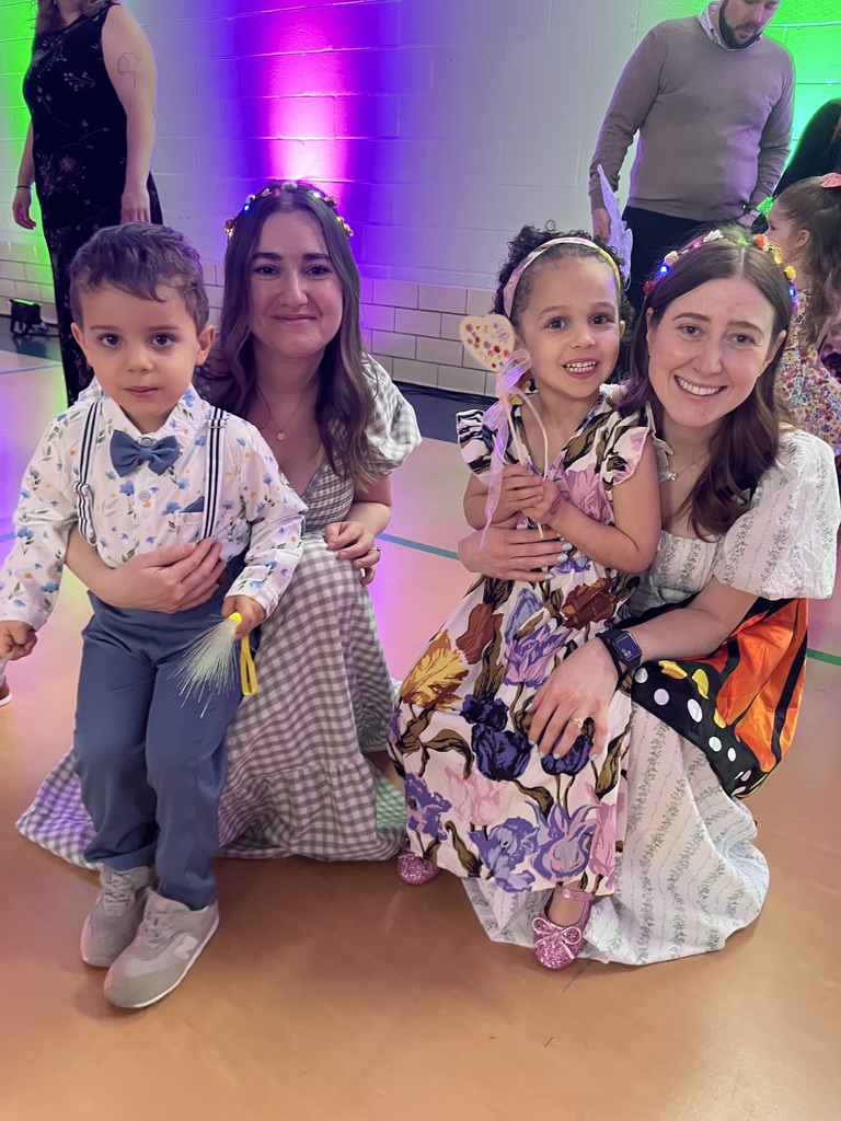 Two teachers and two preschoolers wearing suits and dresses in a gymnasium with party lights, smiling at the camera