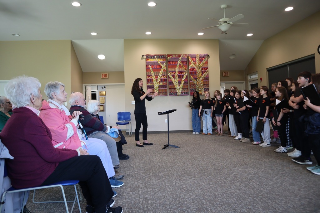 A group of students sing in a choir to a group of Senior Center members