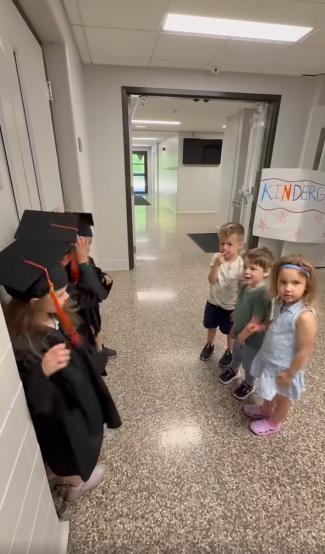 A group of Kindergarteners wearing their black cap and gown stand next to a group of preschoolers