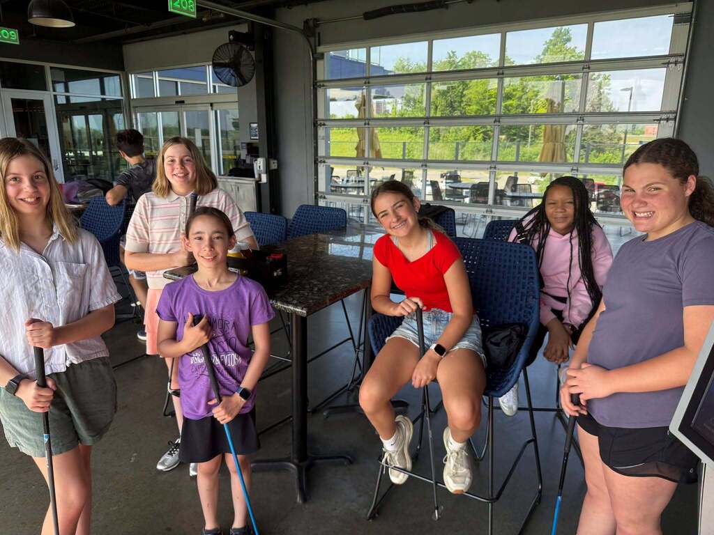 A group of teen campers at TopGolf, holding golf clubs and smiling and the camera