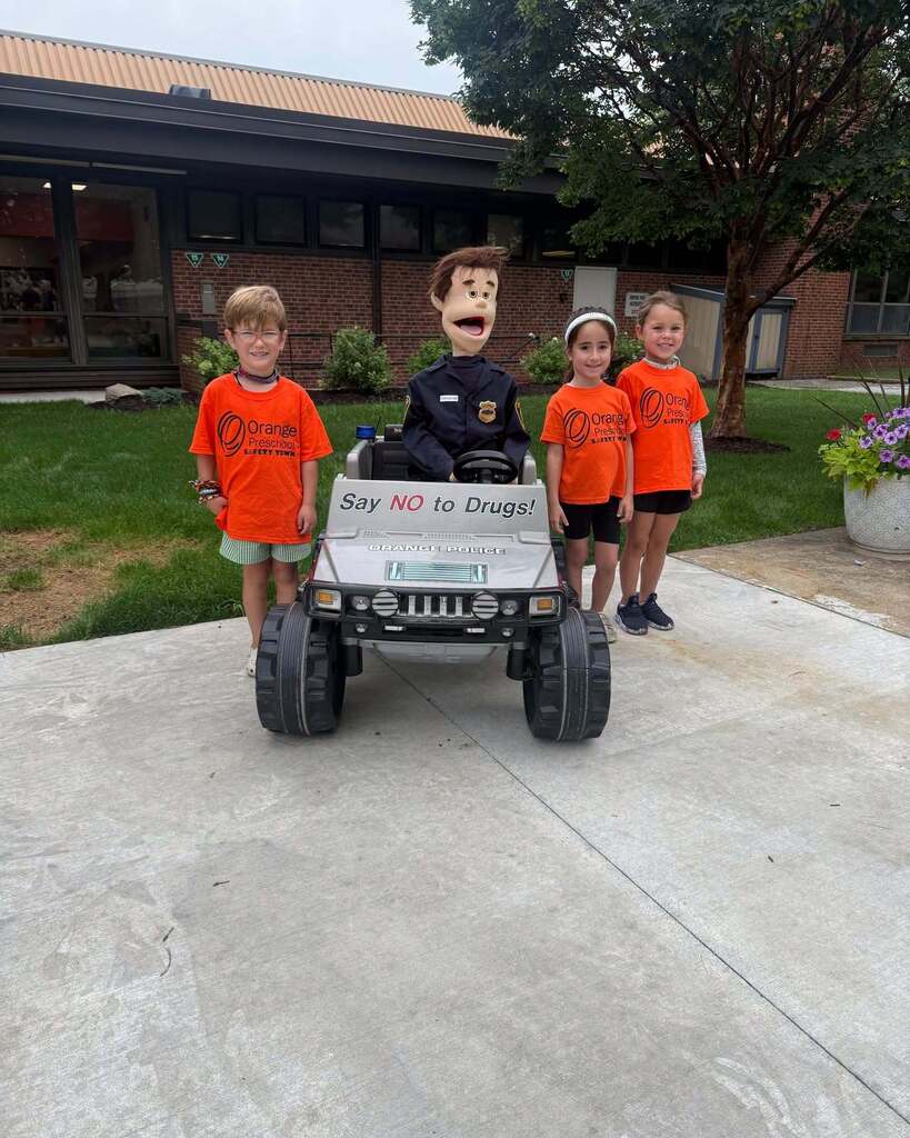 A group of kids stands around a makeshift police car wearing Orange preschool tshirts