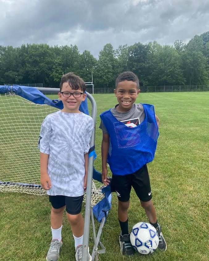 Two boys with a soccer ball, standing in front of a soccer net, smiling at the camera