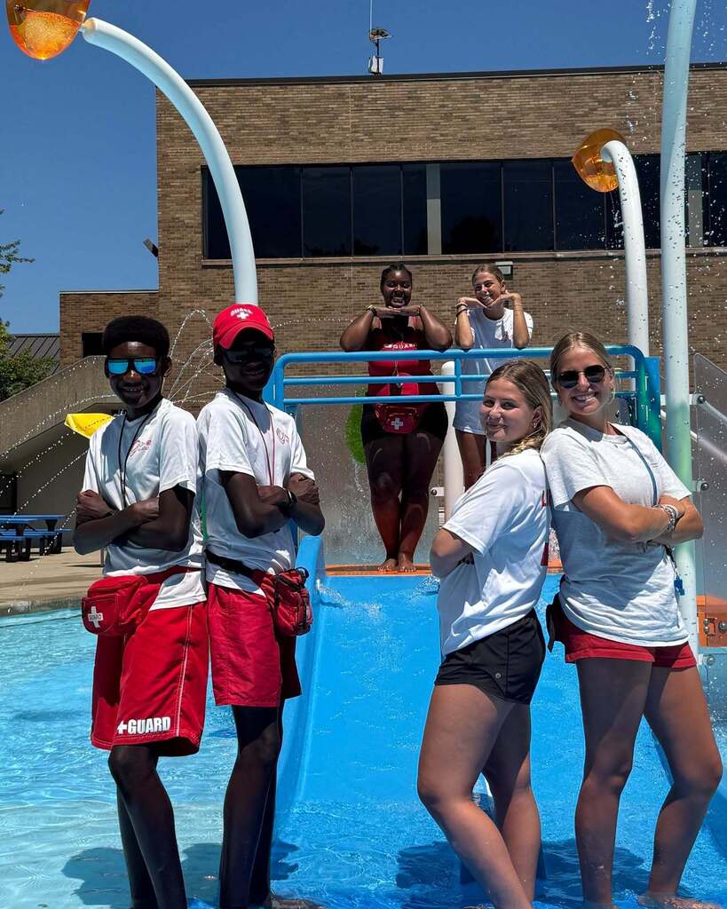 Four lifeguards pose near a water feature in the Orange pool and smile at the camera