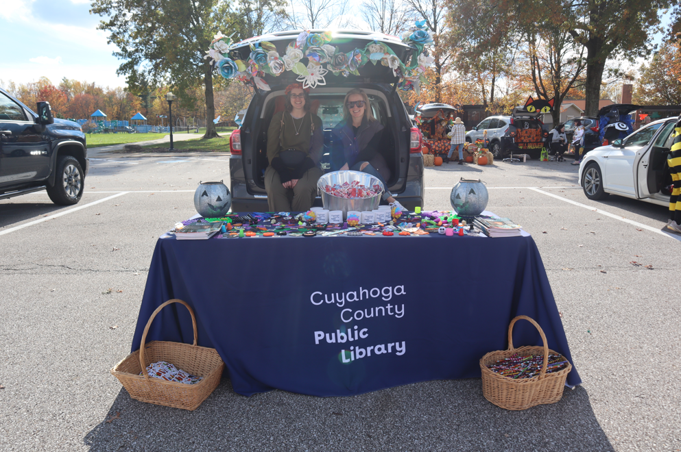 Two people sitting in the back of a car trunk with a "Cuyahoga County Public Library" tablecloth over a table and candy on the table