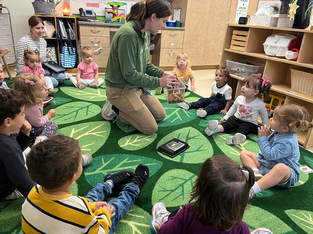 A circle of preschoolers sit on a rug and listen to a teacher speaking in the middle