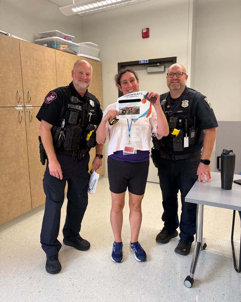 A young woman stands in between two police officers holding up a certificate and smiling at the camera