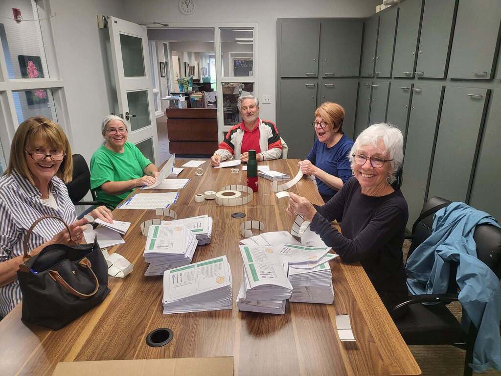 A group of senior citizens sits at a table and stuffs newsletters into envelopes