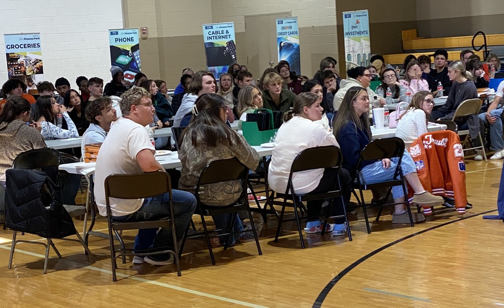 students sitting at tables in a gym listening to a presentation