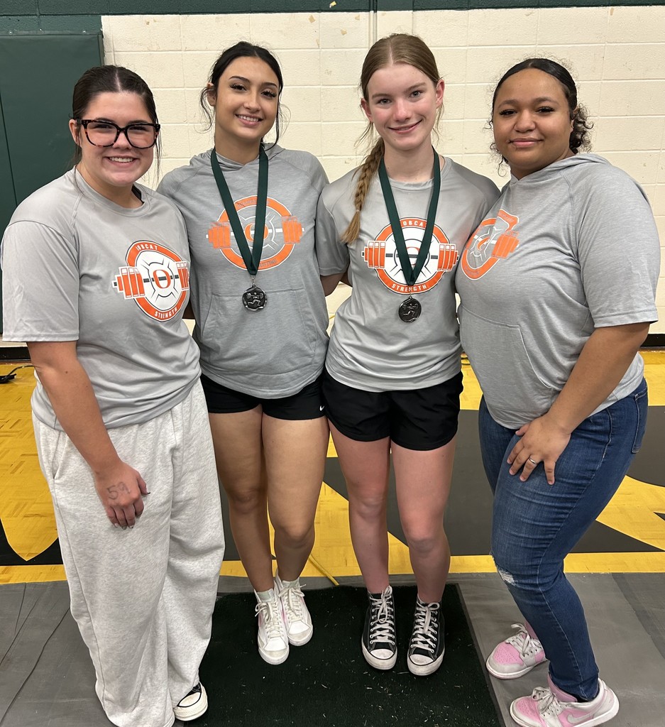Four women in matching gray shirts pose in a gym. Two women wear medals and stand on a mat.