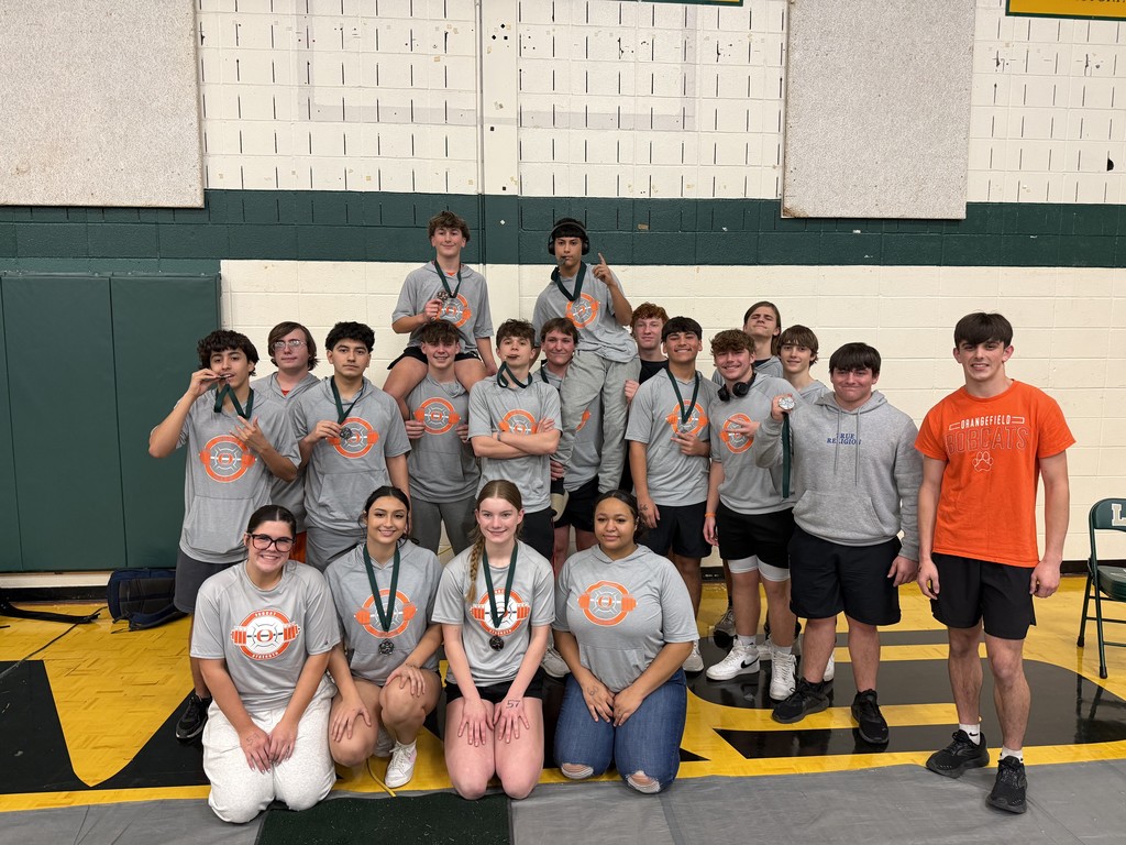 A group of young athletes with medals pose in a gym. They wear matching shirts and stand on a mat.