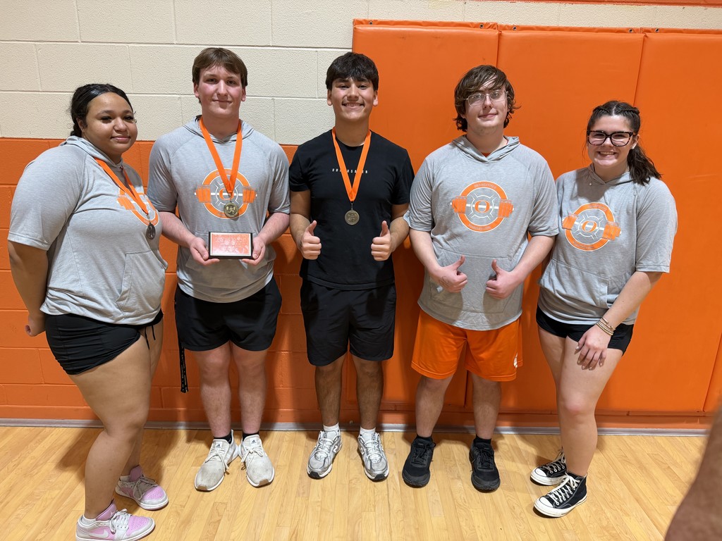 Five people stand together in a gym. They wear matching shirts and shorts. They have medals around their necks. One person holds a plaque. They pose for a photo.