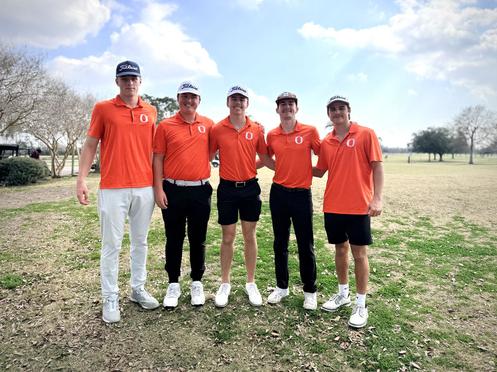 Five men in orange shirts and black pants, standing together on a grassy field, smiling for a photo.