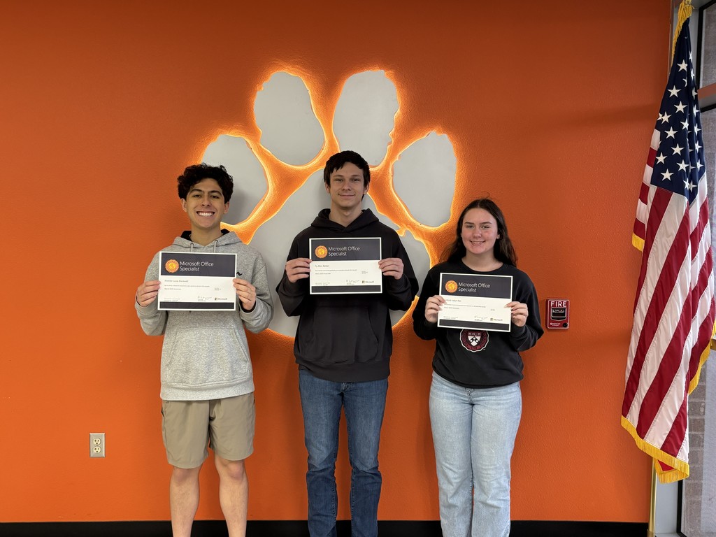 Three students stand in front of an orange wall, holding certificates. The wall features a paw print. An American flag is nearby.