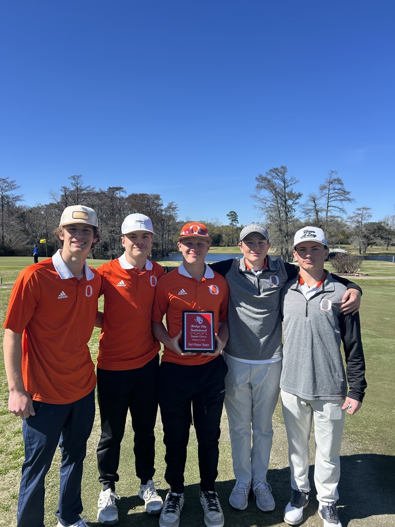 Five people stand on a golf course, holding a plaque. They wear hats and sneakers. Behind them, a clear sky and trees.