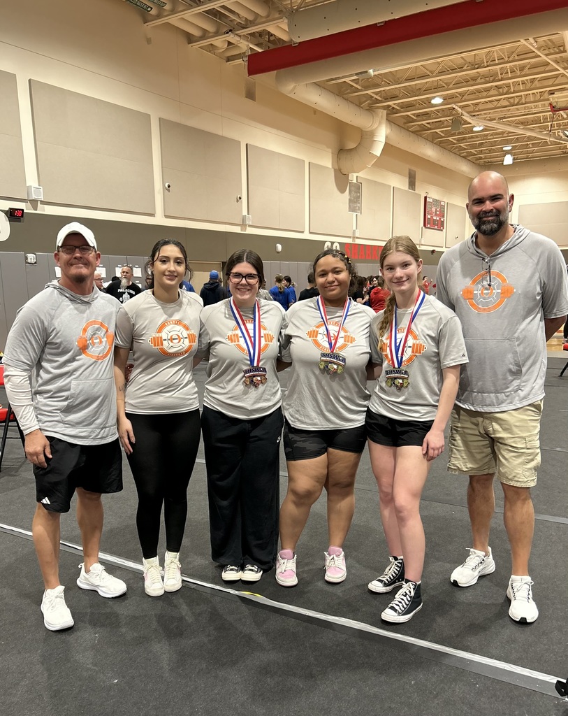 Six people stand together on a gym floor, wearing matching gray shirts and medals. Two women wear black pants, and four have white shoes.