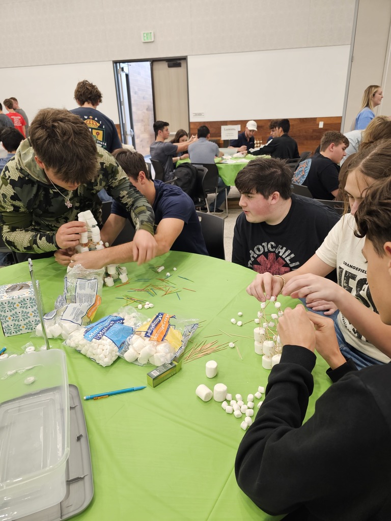 Group of students in a room, seated around a green table, working on a project with marshmallows and sticks.