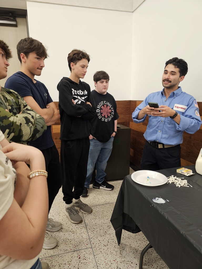 Five boys in a room with a table; one holds a phone, two look at him, one turns away, one watches, and one looks down.