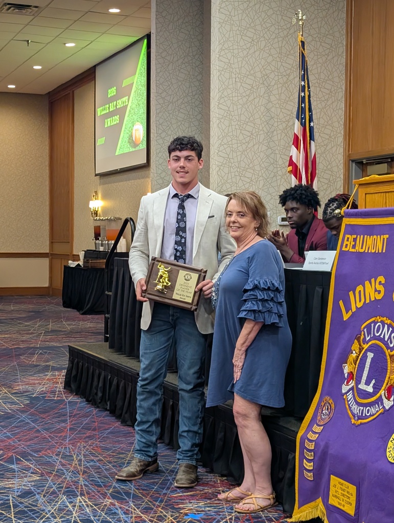 A man in a suit and tie holds an award, standing next to a woman in a blue dress. Behind them, a flag and a purple banner are visible.