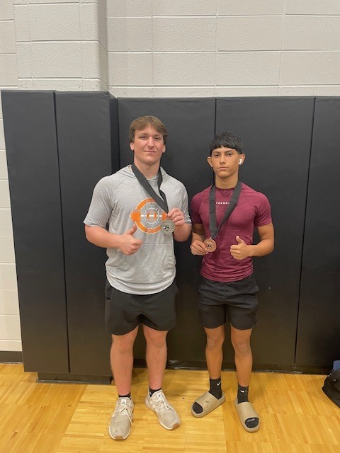 Two men stand on a wooden floor, both wearing medals. One wears a gray shirt, the other a red one. Behind them is a black wall.