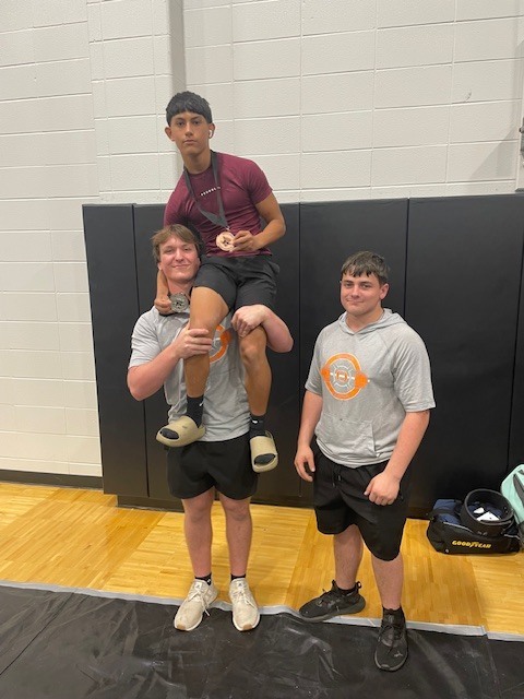Three individuals stand in a gym; one is lifted by two others. They wear matching gray shirts and black shorts.