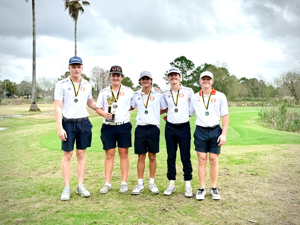 Five individuals stand on a golf course, wearing white shirts, black shorts, and caps. They hold medals and a trophy. Behind them is a grassy field with trees.