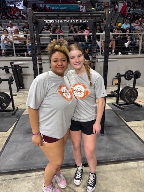 Two women stand before a weight rack in a gym, both wearing gray t-shirts. One wears maroon shorts and the other black.