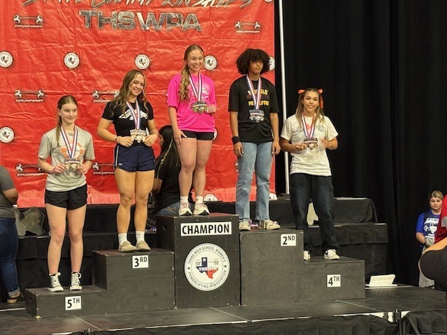 Five women stand on a stage, two on the ground level, three on podiums. They wear medals. Behind them, a banner with the text "TX-WUCA" is displayed.