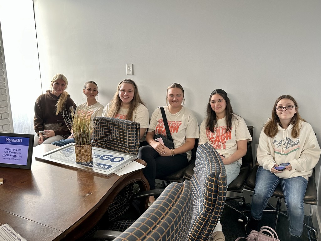 A group of students sitting in a meeting room, some with glasses, wearing white shirts with text.